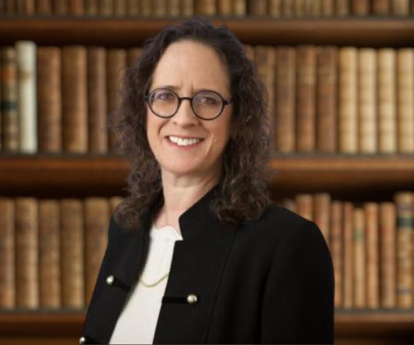 Alyza Lewin, glasses, black jacket, in front of bookshelf bg